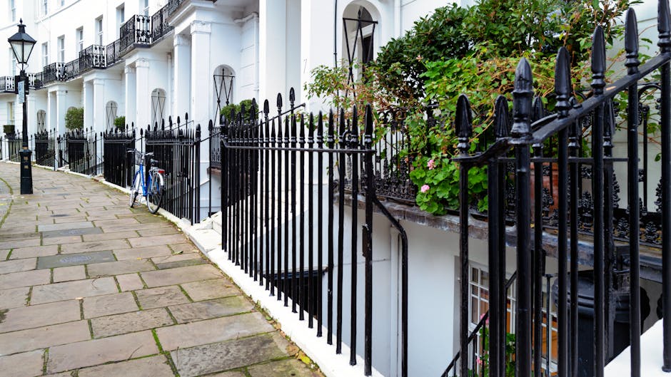 A cobblestone pavement running alongside a row of white terraced houses with black wrought iron railings and gates. The pavement is composed of large, rectangular stones with some moss and dirt in the